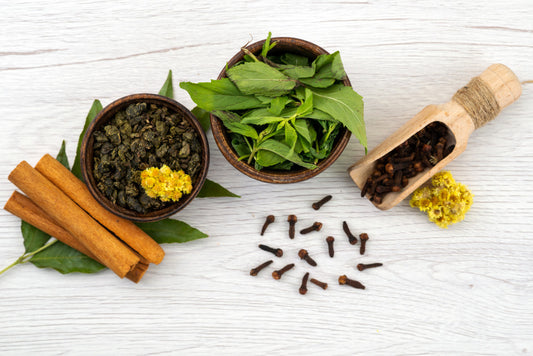 Spices and herbs including cinnamon sticks, cloves, and a bowl of green leaves on a white wooden surface.